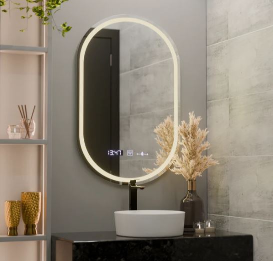 Modern bathroom vanity with a backlit oval mirror showing a digital clock, a white round vessel sink on a dark countertop, and a vase of dried pampas grass nearby.