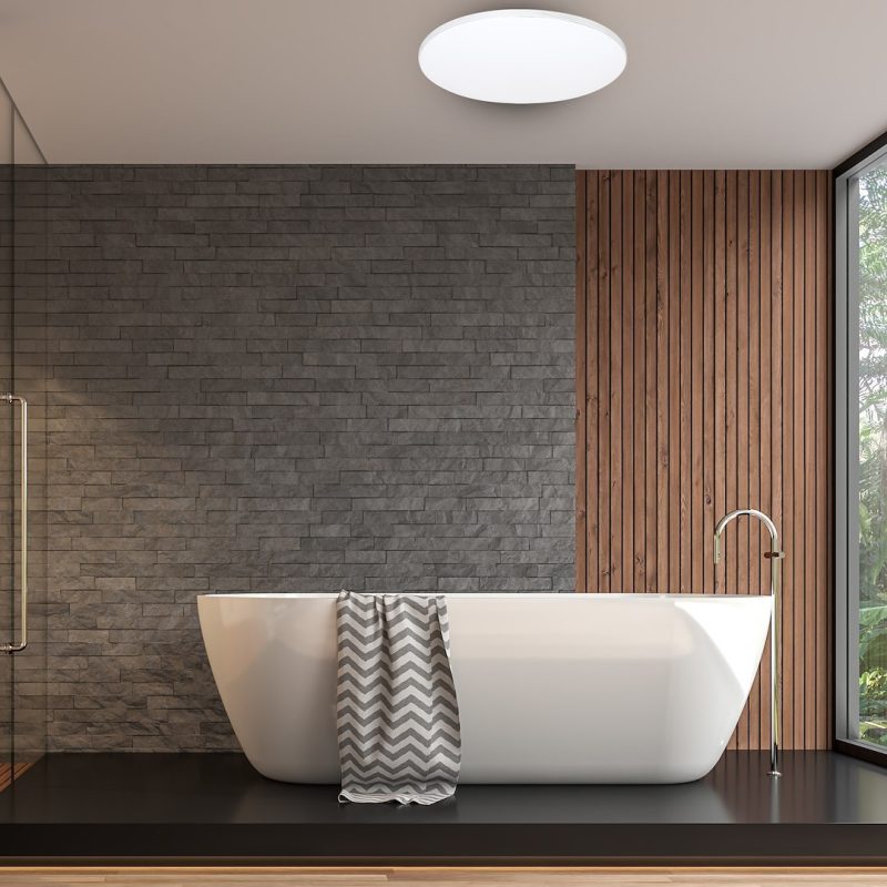 Freestanding white bathtub on a dark platform with a gray zigzag towel, in a modern bathroom with gray stone wall and vertical wood panels.
