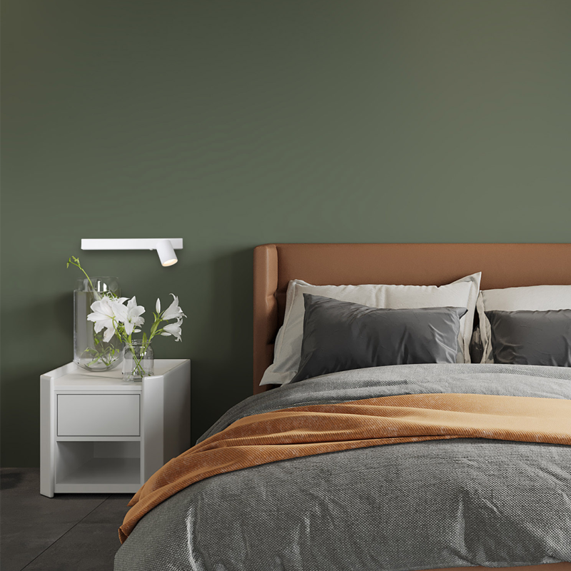 Modern bedroom with an olive-green wall, white nightstand, glass vase of white lilies, wall-mounted lamp, and a bed with gray bedding and an orange throw.