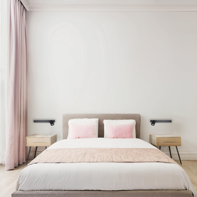 Bed centered in a bright, minimalist bedroom with blush pink pillows, beige headboard, and matching nightstands with black reading lamps on either side.