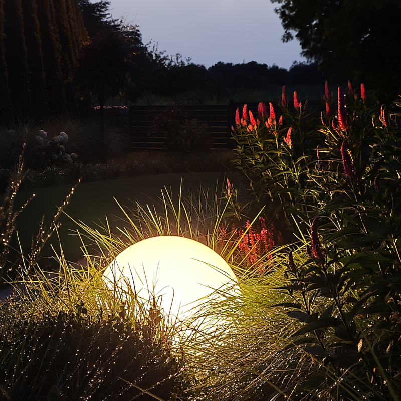 Glowing white sphere light in a dark garden, surrounded by tall grasses and pink-red flowering plants at dusk.