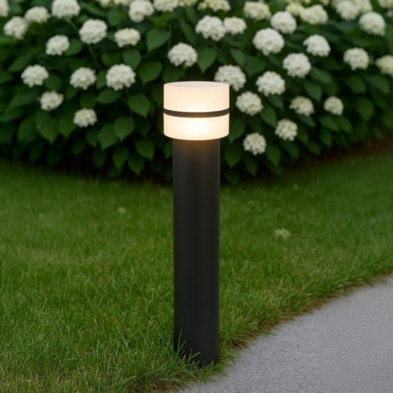 Outdoor path light post glowing in a garden with white hydrangea bushes in the background on a grassy lawn.
