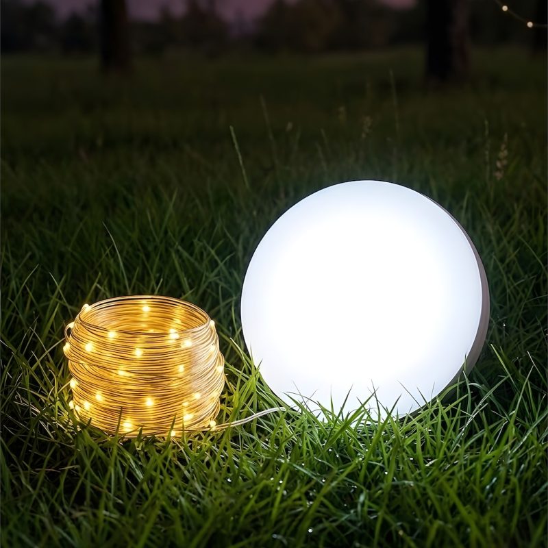 Glowing white sphere light beside a warm string-light coil on grass at dusk.
