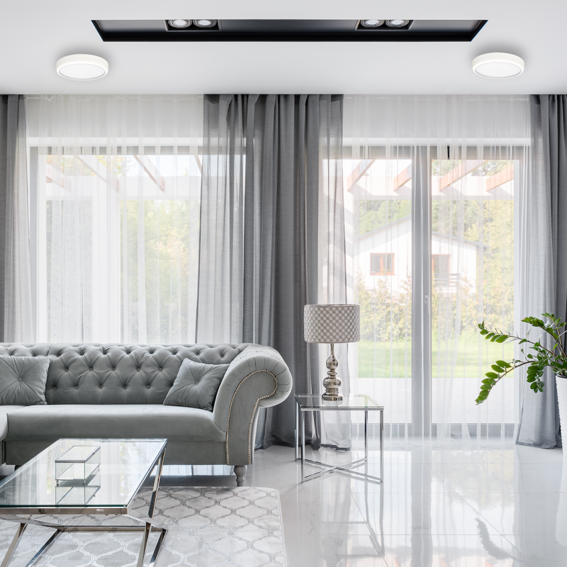 Luxurious living room with a tufted gray sofa, glass coffee table, metal side table with a lamp, sheer and gray curtains, and a potted plant by large windows.