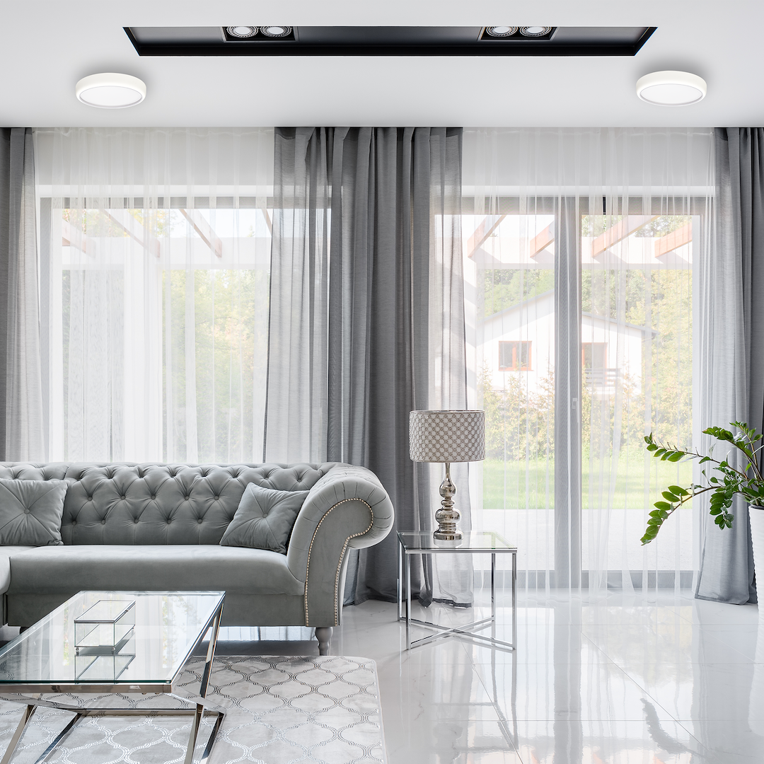 Luxurious living room with a tufted gray sofa, glass coffee table, metal side table with a lamp, sheer and gray curtains, and a potted plant by large windows.