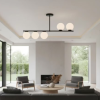 Modern living room with a central fireplace, round coffee table, gray seating, a black rod chandelier with globe lights, and large windows overlooking greenery.