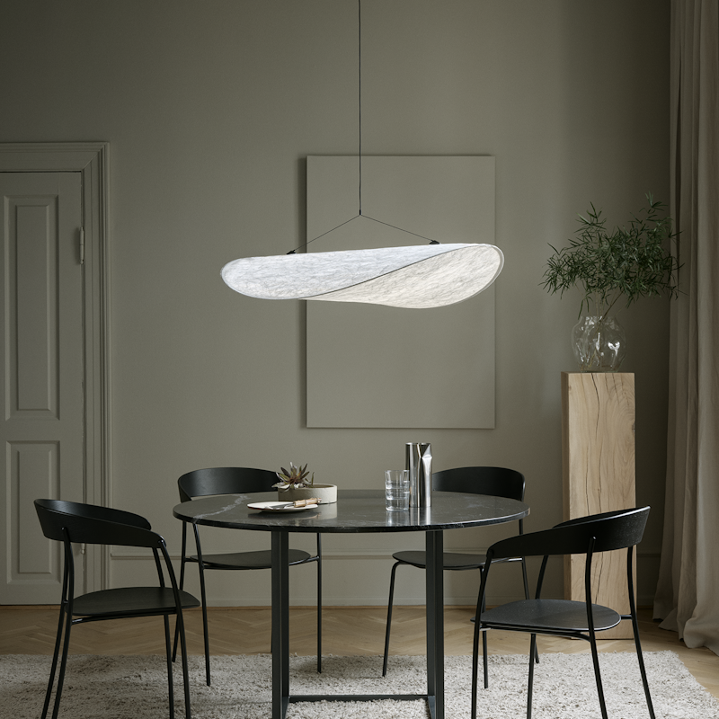 A modern dining room with a round black table, four black chairs, a sculptural hanging light, and a potted plant on a wooden pedestal against a neutral wall.