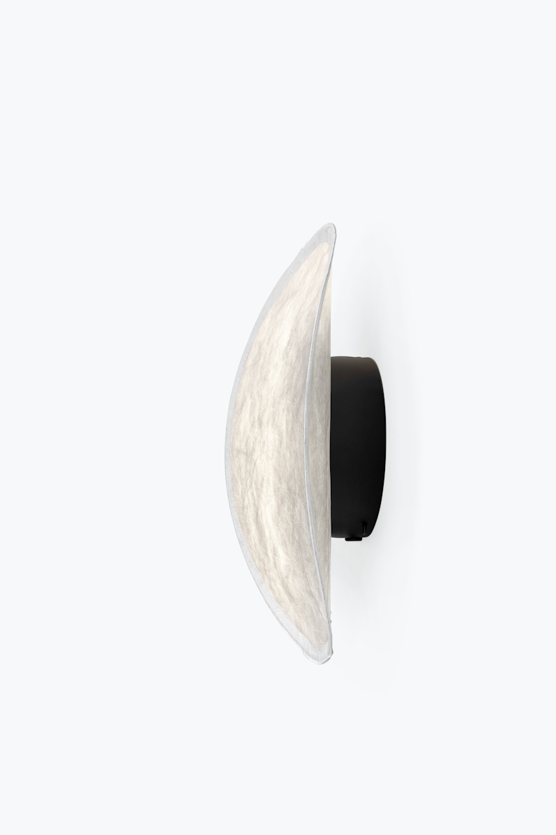 Side view of a wall-mounted circular fabric lampshade attached to a black backing on a white wall.
