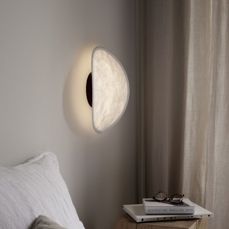 Soft warm wall light above a bedside, with a white pillow and a wooden nightstand holding books and glasses.