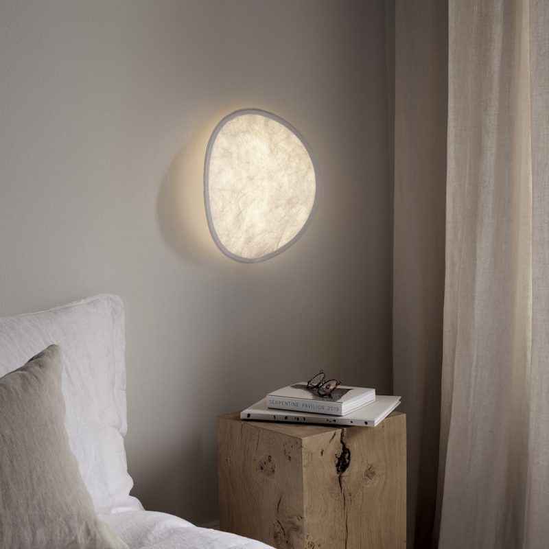 Soft-lit circular wall light above a minimalist wooden nightstand with a stack of books and glasses beside a white bed.