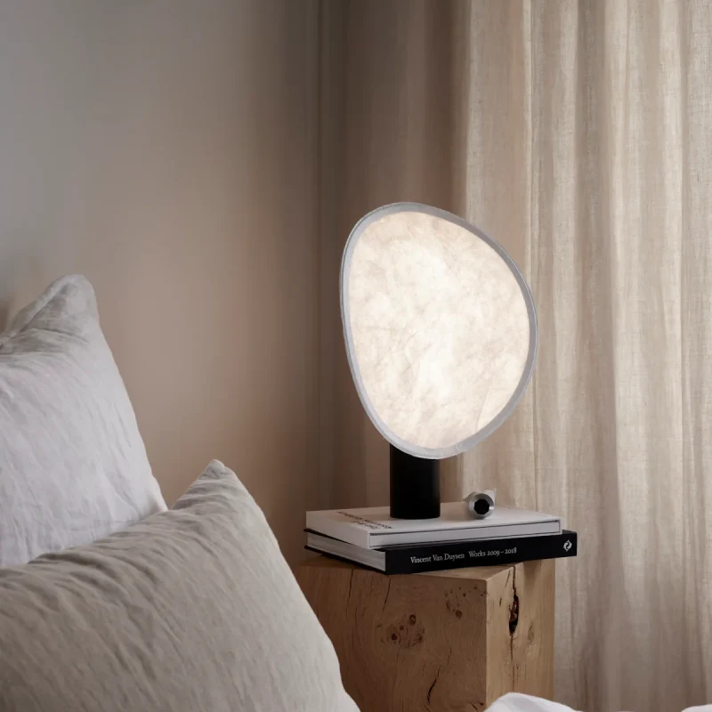 Round fabric lampshade on a black stand, lit softly, perched on a stack of books on a wooden bedside table beside a bed with white linens.