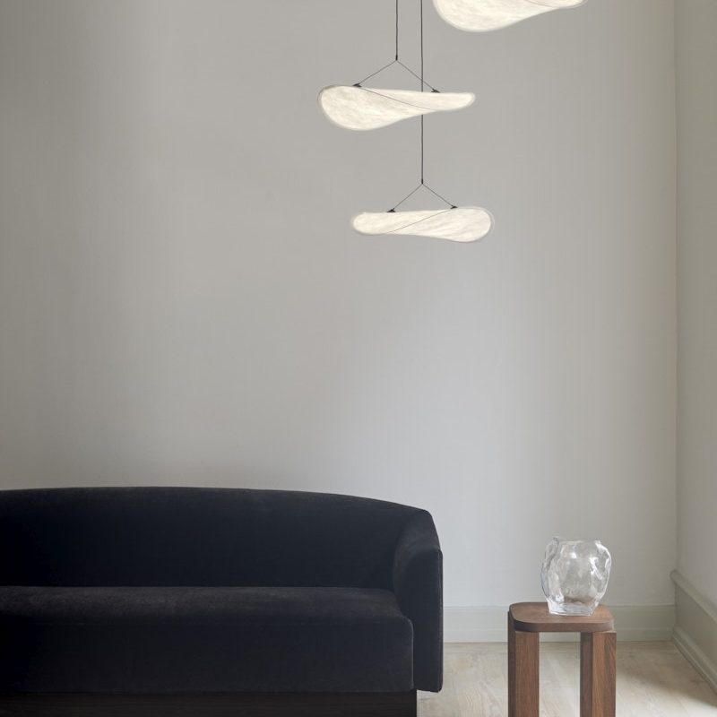 Minimalist living room with a black sofa, light wood floor, and a small wooden side table holding a clear glass vase; sculptural pendant lights hang above.