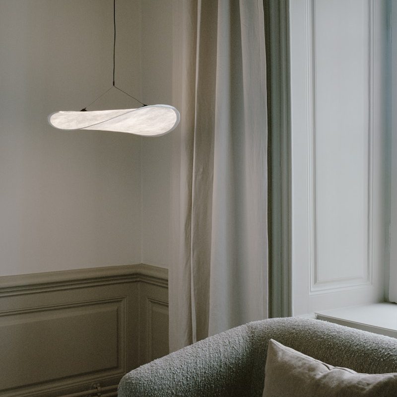 Minimalist hanging light fixture over a gray sofa in a softly lit living room with draped curtains and paneled walls