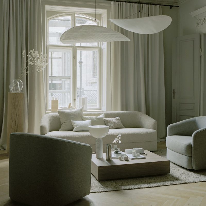 Neutral living room with beige sofa, gray chairs, and a wooden coffee table arranged on a pale rug by a large window with white curtains and modern pendant lights above.