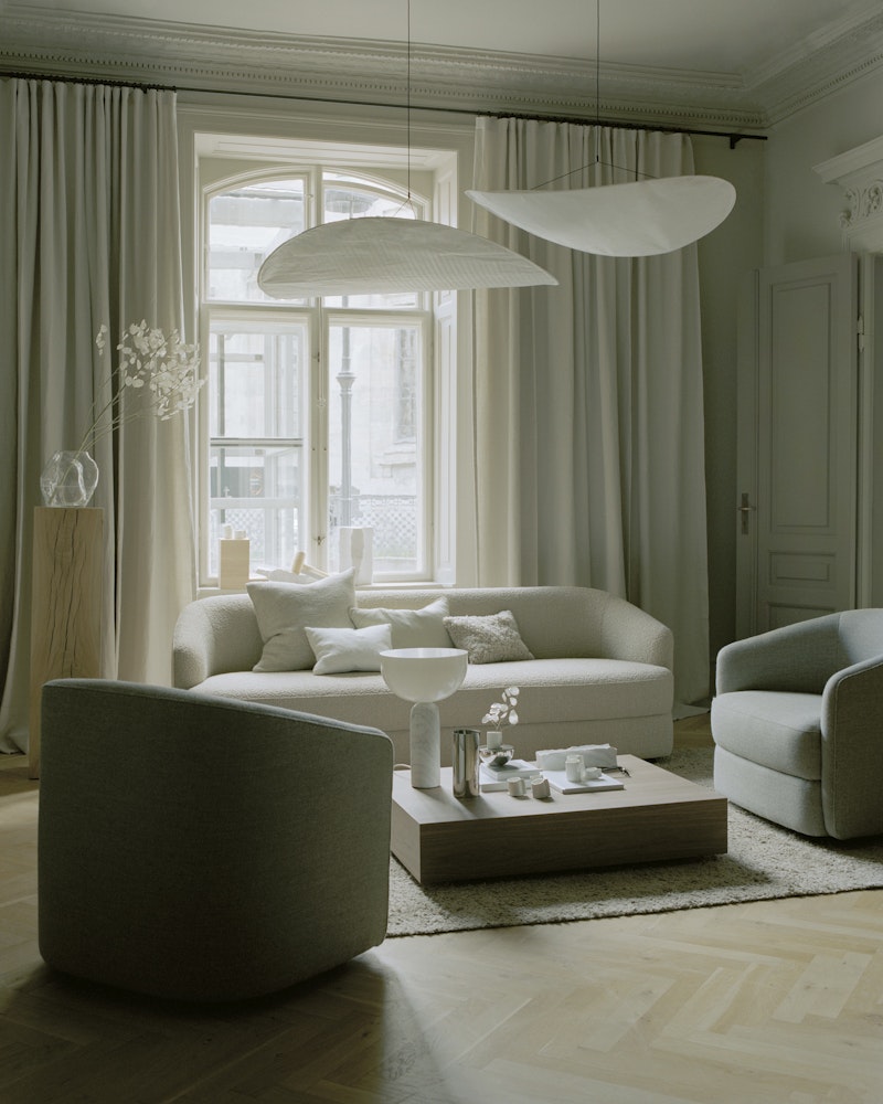 Neutral living room with beige sofa, gray chairs, and a wooden coffee table arranged on a pale rug by a large window with white curtains and modern pendant lights above.