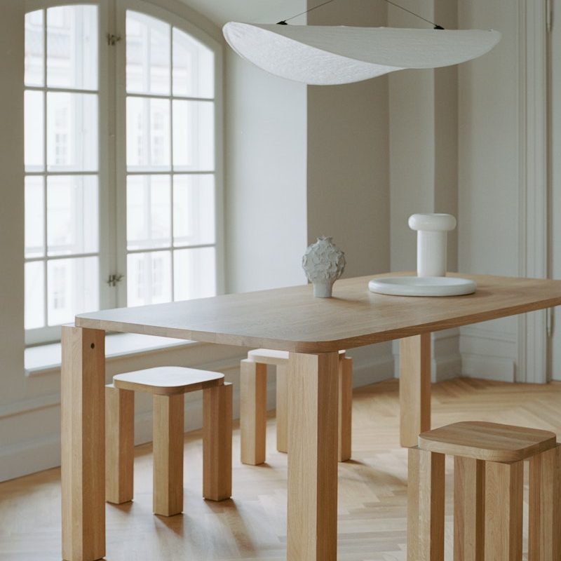Minimalist wooden dining table with matching blocky stools in a bright room, a white pedestal and small sculpture on the tabletop.
