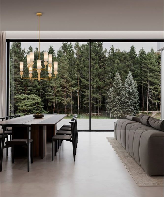 Open-plan dining and living area with a long dining table and black chairs, a gold chandelier, and a gray sectional sofa, framed by floor-to-ceiling glass walls overlooking trees.