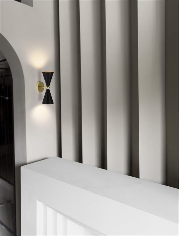 Modern wall sconce with a gold circular mount and two black conical shades on a light gray paneled wall beside a white railing.