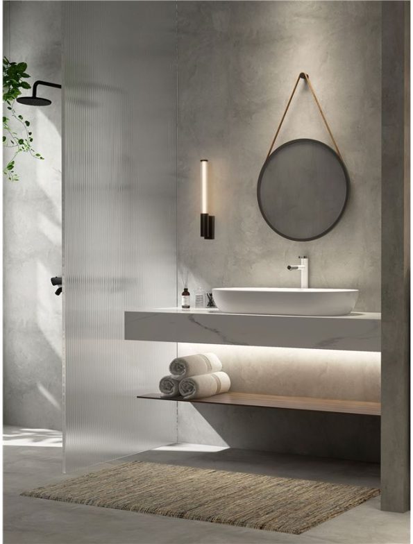 Modern bathroom with round mirror, white vessel sink on a marble counter, stacked towels on a wooden shelf, and a glass shower on the left.