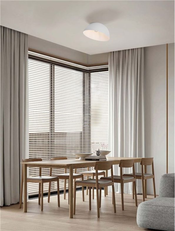 Wooden dining table with six matching chairs in a bright corner, large windows with blinds and white curtains, and a white ceiling light above.
