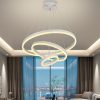 Modern circular ring chandelier with three interlocking illuminated rings hanging from the ceiling of a contemporary living room, large glass windows behind.
