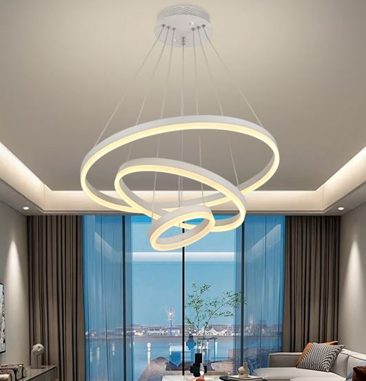 Modern circular ring chandelier with three interlocking illuminated rings hanging from the ceiling of a contemporary living room, large glass windows behind.