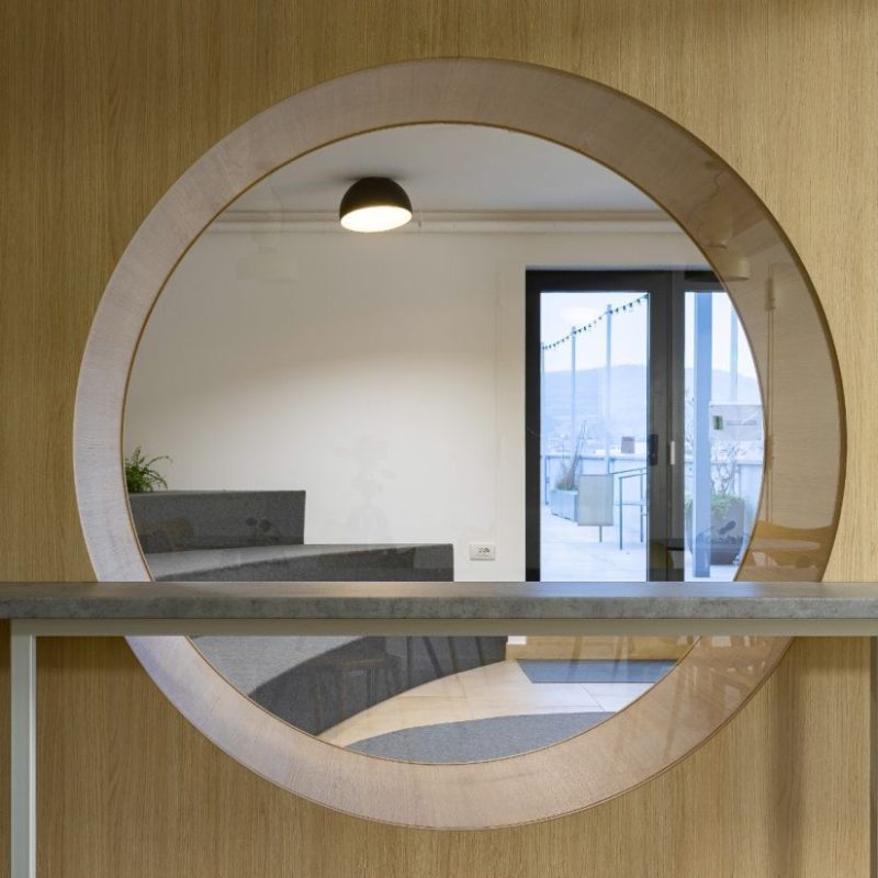 Round circular mirror in a wood-paneled wall reflecting a seating area and an outdoor patio door.