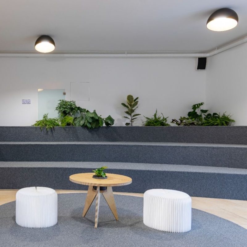 Modern indoor lounge with tiered gray platforms, a round wooden table, two white ribbed stools, and potted plants along a white wall with ceiling lights.