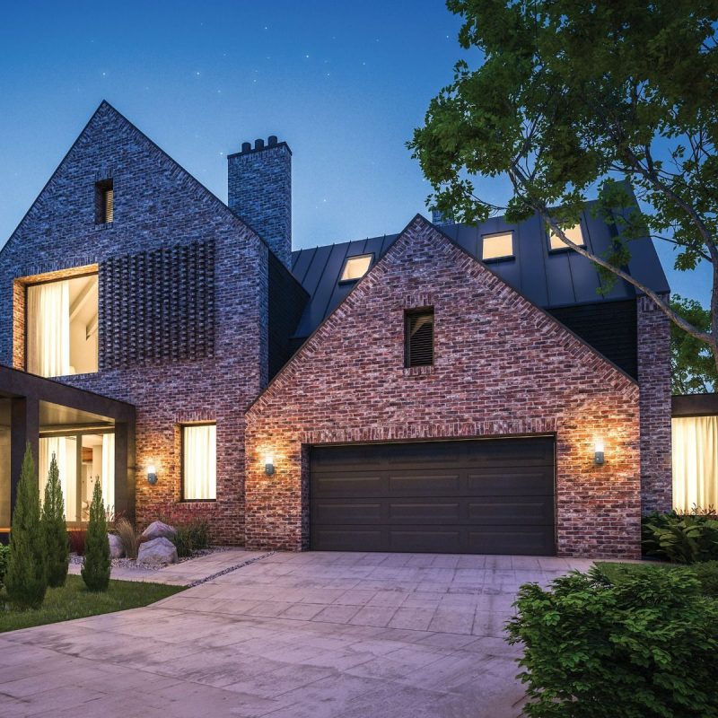 Two-story brick house with a dark garage door, illuminated entry lights, and a stone driveway at dusk.