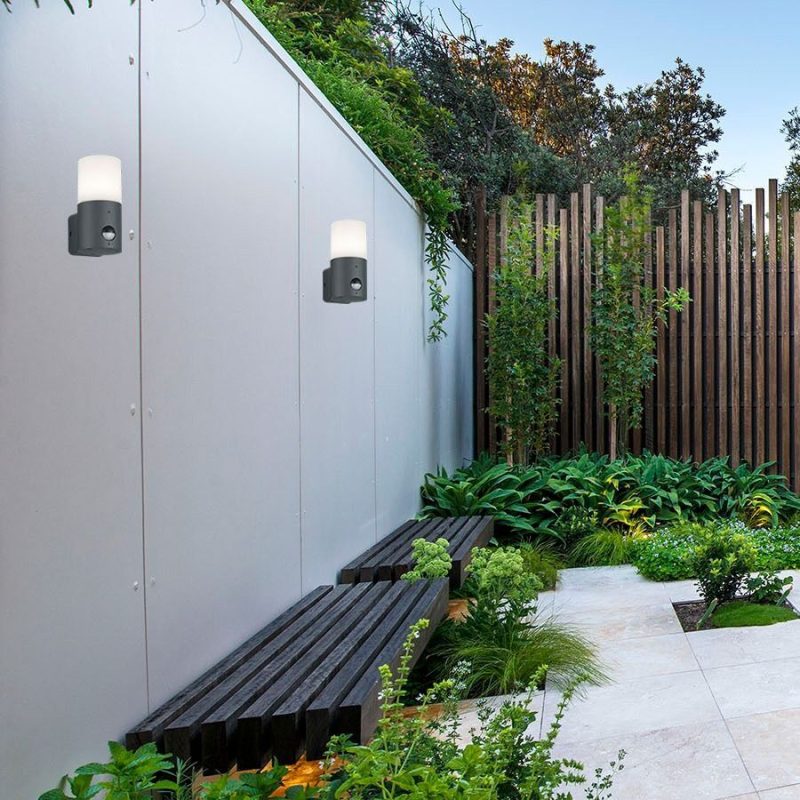 Contemporary courtyard with a gray metal wall, black slatted benches, and wall-mounted lights amid lush greenery and a wooden fence.